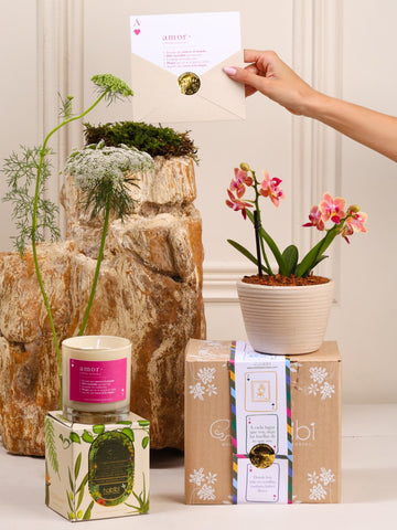 Kit True Love: orquídea baby sembrada en matera Sahara de barro blanco con relieve, junto a la vela Cupido y la postal de Amor, sobre fondo de la temporada Cartas de Amor.